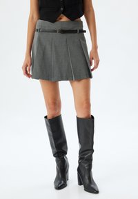 Falda corta gris plisada con un cinturón negro, combinada con botas altas de cuero negro con tacones. Textura suave, diseño a medida y hasta la rodilla.