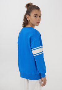 Felpa blu con strisce bianche e azzurro chiaro sulle maniche, polsini a costine e collo a giro. Il tessuto sembra morbido con una vestibilità casual.