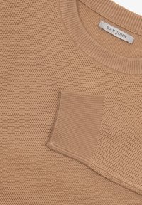 Strikpullover i lys beige, med et struktureret honningcomb-design og en brystlomme. Ribkantet halsudskæring og manchetter giver struktur.