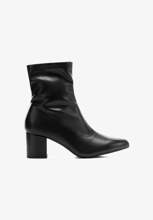 Bota de tobillo de cuero negro con tacón grueso y puntera puntiaguda, parte superior lisa con costuras sutiles, fotografiada sobre fondo blanco.