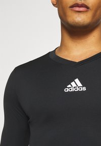 Schwarzes langärmliges Sportshirt mit V-Ausschnitt, aus strukturiertem Stoff und mit einem weißen Adidas-Logo auf der Brust.
