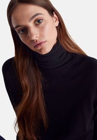 Černý rolák se žebrovanou texturou, přiléhavý střih a vysoký límec. Dlouhé hnědé vlasy částečně viditelné podél výstřihu.