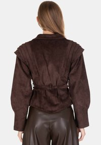 Giacca marrone in finta pelle scamosciata con spalle plissettate e vita con cintura. Presenta maniche lunghe e un design posteriore strutturato e allungato.