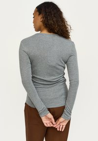 Šedé elastické tričko s dlouhým rukávem, žebrovanou strukturou a kulatým výstřihem, doplněné volnými hnědými kalhotami.