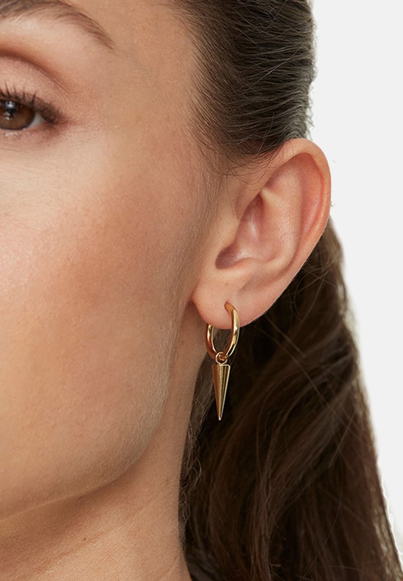 Gros plan sur l’oreille d’une femme aux cheveux foncés montrant une boucle d’oreille en or en forme de cercle avec un pendentif conique pointu.