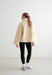 Beige fleecová bunda s vysokým límcem, širokými rukávy a texturou, spárovaná s černými legínami a sportovními botami.
