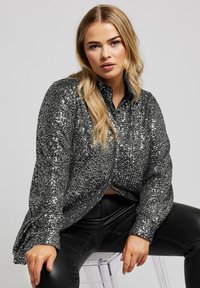 Blusa nera in paillettes con colletto a camicia, maniche lunghe e vestibilità rilassata, abbinata a pantaloni di pelle nera. Texture lucida e trasparente.