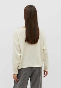 Cremfarbener Pullover mit lässigem Schnitt, weitem Halsausschnitt und langen Ärmeln, kombiniert mit grauen Nadelstreifenhosen. Weiche Textur und minimalistisches Design.