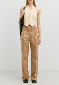 Beige cropped gilet met knopen, gecombineerd met lichtbruine op maat gemaakte broek. Het model houdt een zwarte oversized handtas vast. Glad stof en gestructureerde pasvorm.