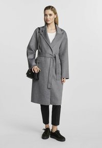 Szary wełniany płaszcz z paskiem w talii, z kołnierzem typu notch oraz bocznymi kieszeniami. Nosi się go na białej bluzce i czarnych spodniach, do których dobrano czarne snickersy.