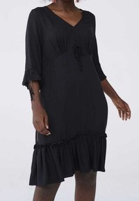 Svart klänning med V-ringning, veckad byst och knytdetalj. Har 3/4 ärmar med volangkant och en flerlagers volangkant nertill. Mjukt tyg.