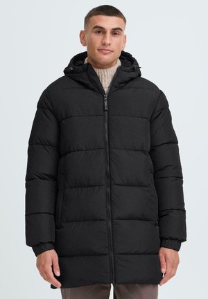 Schwarze aufblasbare Jacke mit Kapuze, ausgestattet mit einem Reißverschluss, gestepptem Design und elastischen Bündchen. Modell trägt einen cremefarbenen Pullover darunter.