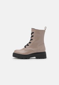Stivaletti in pelle beige con finitura lucida, lacci neri e suola in gomma nera spessa. Caratterizzati da un pratico tirante sul retro.
