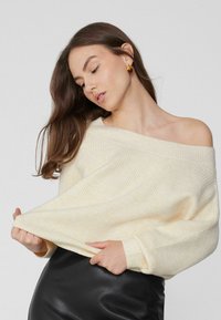 Cremefarbener und hellgelber gestreifter Strickpullover mit einem schulterfreien Design. Lange Ärmel und gerippte Bündchen. Kombiniert mit einem schwarzen Lederrock.