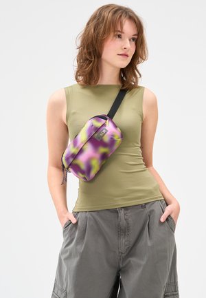 Junge Frau, die ein olivfarbenes ärmelloses Oberteil und lockere graue Hosen trägt, dazu eine lila-grüne Umhängetasche, Hände in den Taschen, schaut nach rechts.