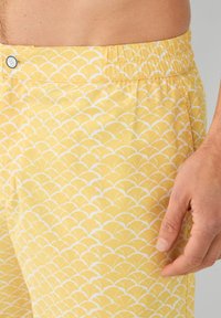 Žute kupaće hlače izrađene od lagane tkanine, s bijelim uzorkom školjki, elastičnim pojasom i gumbom na struku.
