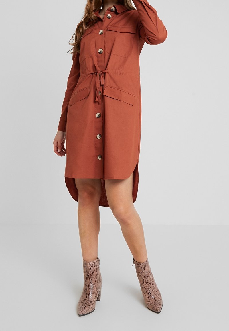Femme portant une robe boutonnée couleur rouille à manches longues et taille à cordon, associée à des bottines à motif peau de serpent.
