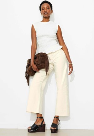 Femme portant un top blanc sans manches, pantalon crème à jambes larges, sandales à plateforme noires, tenant un sac en suède marron à franges contre un mur blanc.