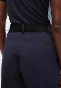 Άτομο που φοράει σκούρο μπλε αθλητικό παντελόνι με μαύρη ζώνη και σκούρο μπλε πουκάμισο κρυμμένο μέσα στο παντελόνι, δείχνοντας μια τσέπη με φερμουάρ στην πλάτη.