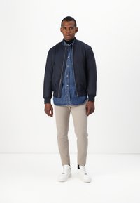 Marine bomberjakke over en denimskjorte, beige bukser og hvite sneakers. Jevn, lett materiale; tettsittende design; ribbestrikkede mansjetter og hem.