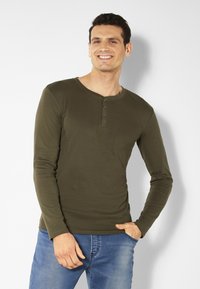 Olivgrünes Langarmshirt mit Rundhalsausschnitt und Drei-Knopf-Leiste, kombiniert mit hellblauen Jeans. Glatte Stoffstruktur.