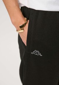 Arm med gullarmbånd hviler i bukselomme med svarte bukser som har hvit Kappa-logo med to sittende figurer rygg mot rygg.