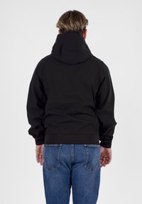 Giacca nera con cappuccio, dotata di polsini e orlo elasticizzati, caratterizzata da una superficie liscia, indossata sopra jeans in denim blu. Vista posteriore mostrata.