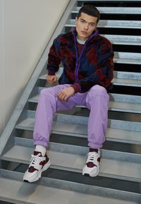 Fleece jack in marineblauw met een rood bloemenpatroon, gecombineerd met lichtpaarse joggers en witte sneakers met rode accenten, zittend op stalen trappen.