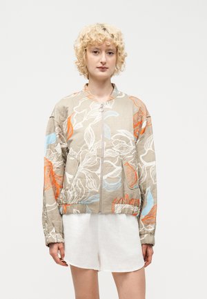 Szőke személy, aki bézs virágmintás cipzáras dzsekit visel narancssárga és kék részletekkel, fehér rövidnadrággal párosítva, semleges háttér előtt áll.