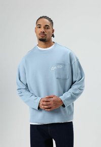 Lichtblauwe sweatshirt, losse pasvorm, met een borstzak en geborduurde details. Draagt over een wit shirt, gecombineerd met donkere jeans.