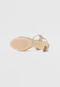 Beige høyhælte sandaler med teksturert overdel, ankelrem og lukket tå. Gummisåle med horisontale spor for bedre grep.