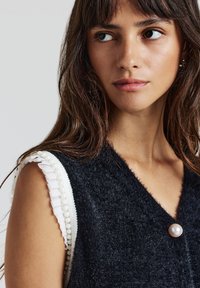 Gilet nero con texture e scollatura a V profonda, arricchito da dettagli in bianco con volant e un bottone a perla. Vista ravvicinata dei dettagli sulle spalle.