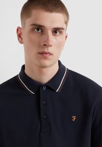 Navy Poloshirt aus Baumwolle, mit einem kragen in braun und weißem Rand, Knopfleiste und einem bestickten orangen Logo auf der Brust.