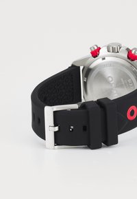 Black rubber watch strap with holes, stainless steel buckle, and polished silver case. Features red accents on the crown and buttons.