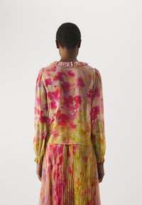 Blusa colorata e trasparente con maniche lunghe e colletto arricciato, caratterizzata da un motivo ad acquerello in tonalità di rosa, giallo e verde.