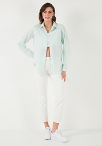Light mint green button-up shirt with a front pocket, paired with white cropped pants and white sneakers. The shirt features a relaxed fit.