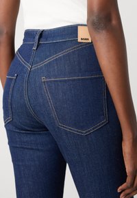 Mørkeblå denimjeans med høy midje, kontrasterende gullsting og to baklommer med et logo-merke ved midjen.