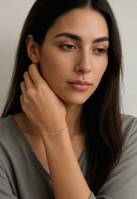 Giovane donna con lunghi capelli scuri che indossa un delicato bracciale a forma di foglia d'argento, mentre si accarezza delicatamente il collo, vestita con una maglia grigia.