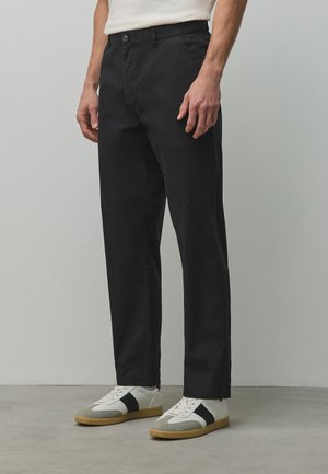 Homme portant un pantalon noir droit associé à des baskets blanches avec des rayures noires et des semelles en gomme, debout sur un sol et un mur gris.