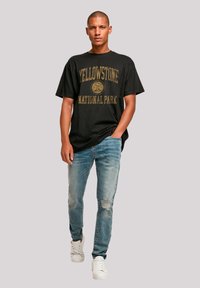 Black cotton t-shirt featuring "YELLOWSTONE NATIONAL PARK" in gold text, paired with light blue distressed jeans and white sneakers.
