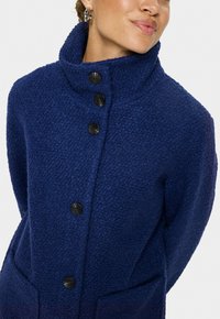 Marinblå rock av texturerad tyg, med en hög krage, flera svarta knappar och två framfickor.