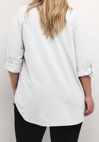 Hvid langærmet skjorte med knapper, opkuffedede ærmer og afrundet nederkant, lavet af letvægtsstof, vist fra bagsiden.