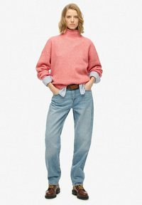 Vrouw staat met handen in haar zakken, gekleed in een roze coltrui, gestreepte shirt, lichtblauwe spijkerbroek en bruine laarzen.