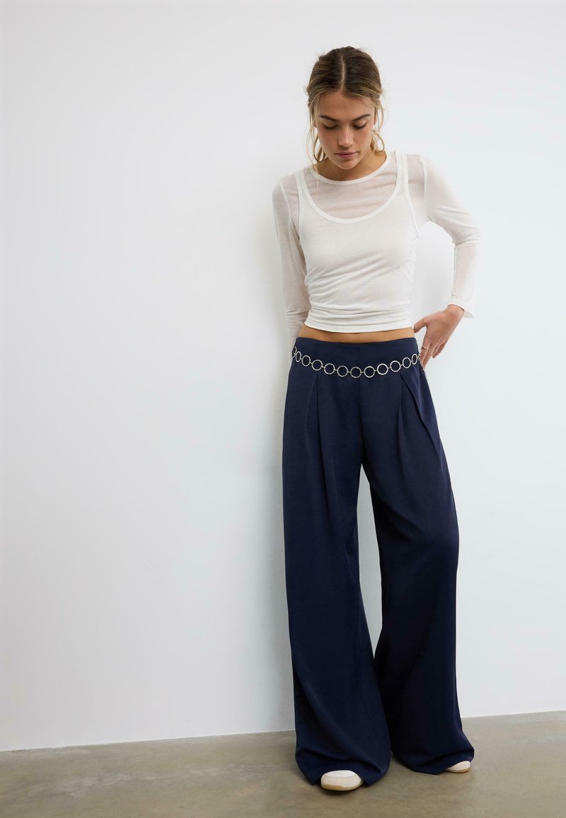Femme portant un haut blanc transparent à manches longues, un pantalon large bleu marine avec une ceinture chaîne argentée, debout contre un mur blanc uni.