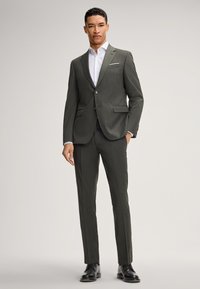 Mężczyzna w dopasowanym szarym garniturze, białej koszuli w paski i czarnych skórzanych butach wizytowych, stoi na tle jasnego, gładkiego tła.