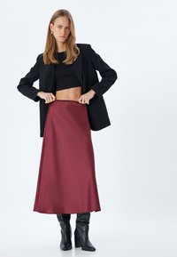 Gonna maxi in satin bordeaux con una texture liscia, abbinata a un top corto nero e una giacca, completata da stivali neri al ginocchio.