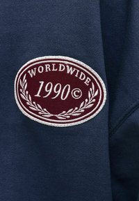 Μπορντό οβάλ πατς σε ναυτικό μπλε ύφασμα με λευκή κεντημένη γραμματοσειρά "WORLDWIDE 1990©" και σχέδια κλαδιών δάφνης γύρω από το κείμενο.