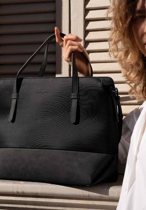 Black tote bag with a textured surface, featuring two sturdy handles and a smooth, darker base. Zipper closure with subtle branding visible.