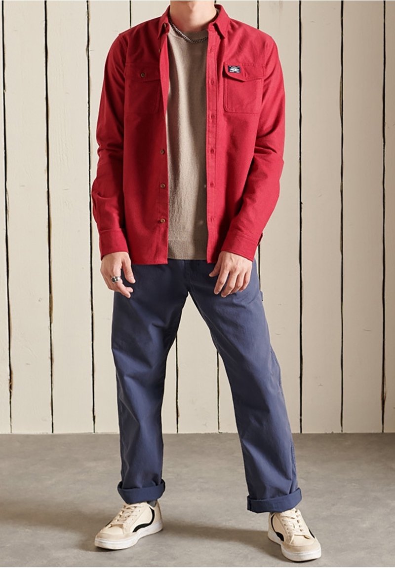 Chemise rouge à boutons portée sur un t-shirt beige à col rond, associée à un pantalon bleu marine retroussé et des baskets blanches, sur un fond en bois.