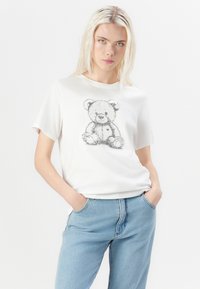 T-shirt en coton blanc avec un imprimé ours en peluche en noir et gris. Manches courtes, col rond, coupe décontractée, associé à un jean bleu clair.